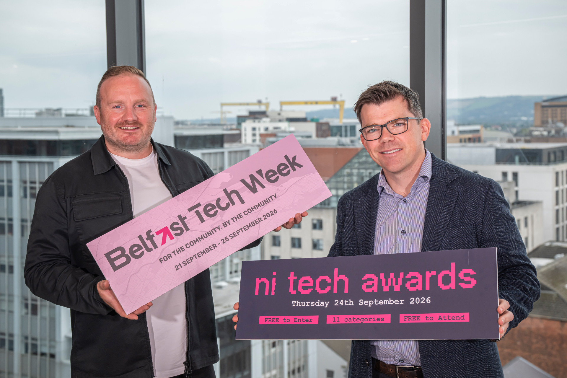 Simon Bailie of Belfast Tech Week and Sean Devlin of MCS Group holding Belfast Tech Week and NI Tech Awards signs above the Belfast skyline